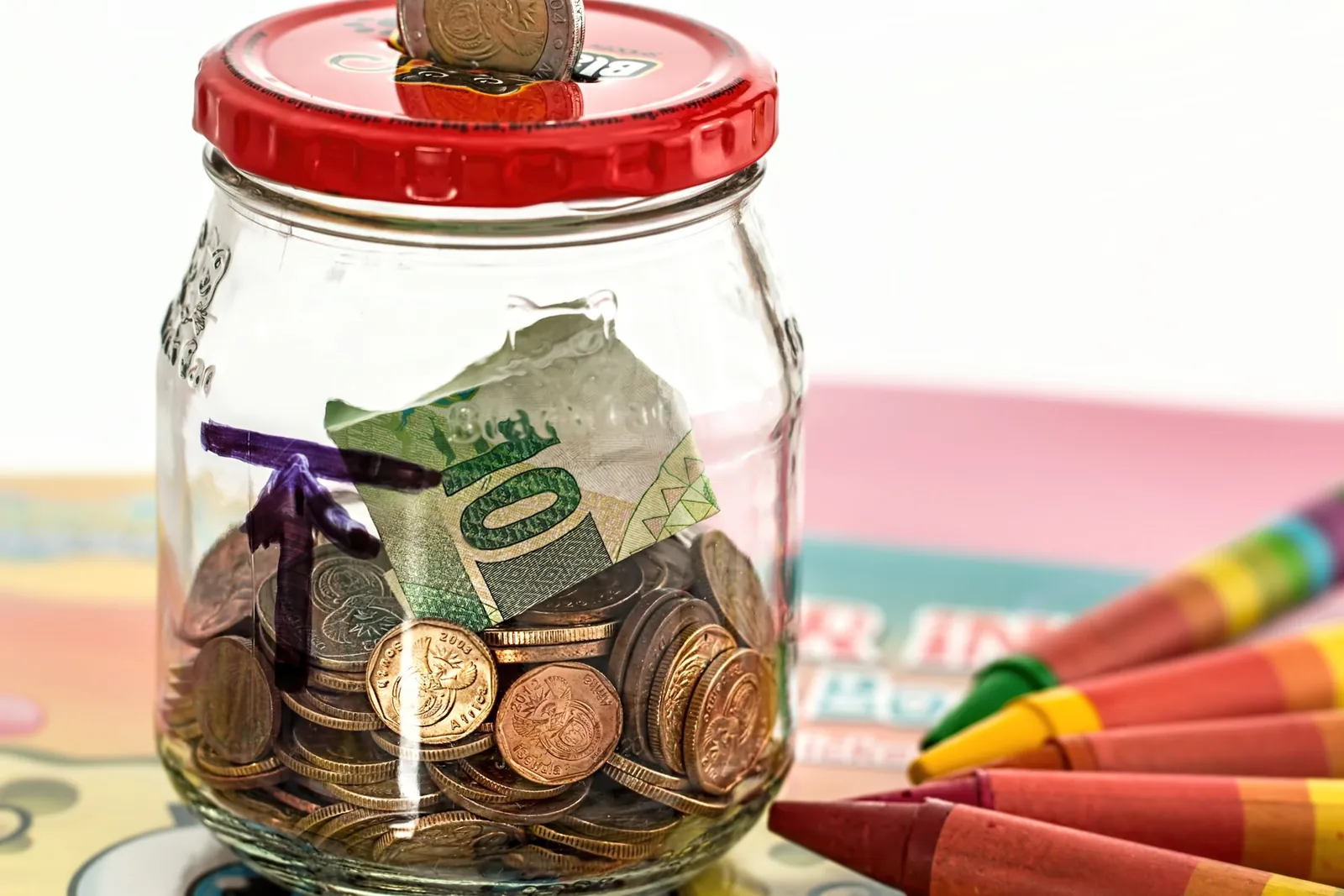 A glass jar with a red lid stuffed with coins and a folded banknote, next to a row of crayons