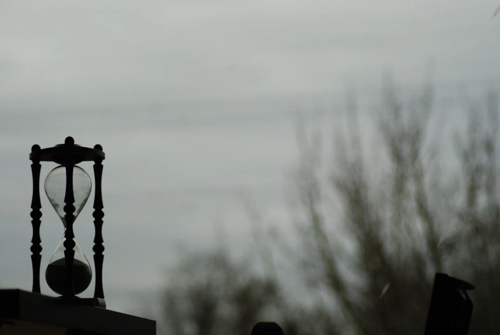 Silhouette of an hourglass on a ledge against a gray winter sky with bare trees in the background
