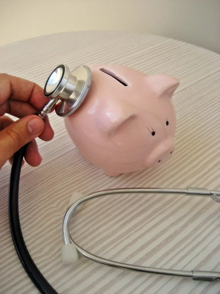 A hand holding a stethoscope against a pink ceramic piggy bank on a striped tabletop