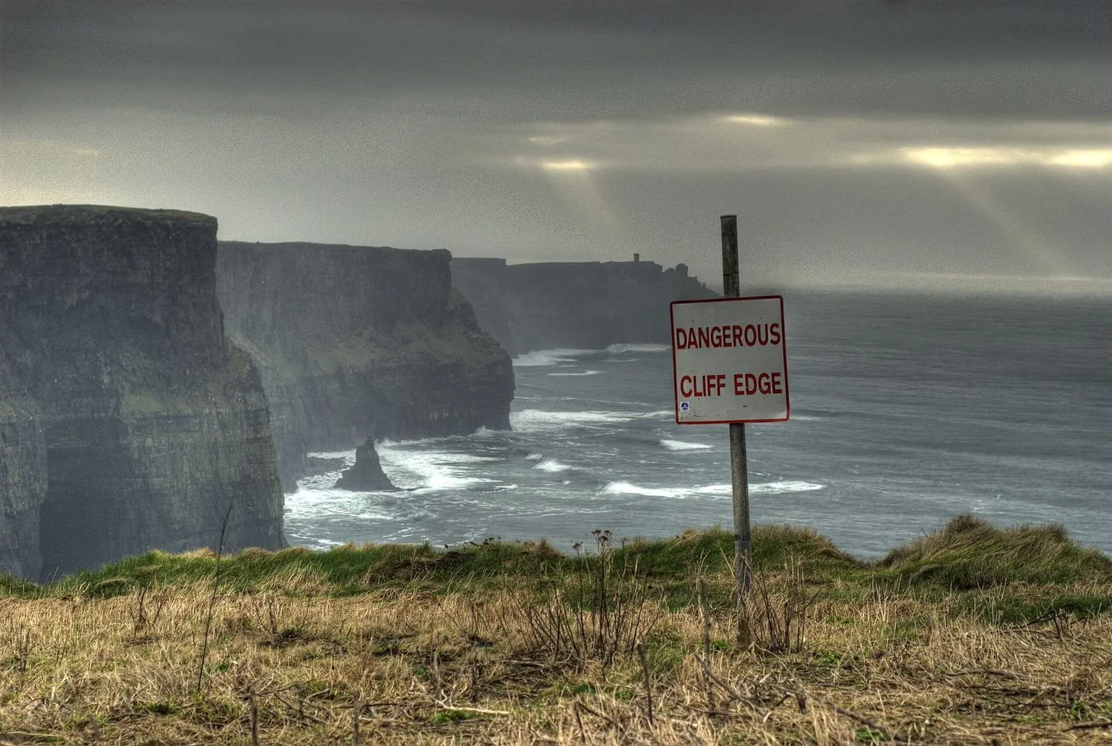 Red-and-white "Dangerous Cliff Edge" sign on a grassy clifftop above dark sea cliffs and crashing waves under a stormy sky.