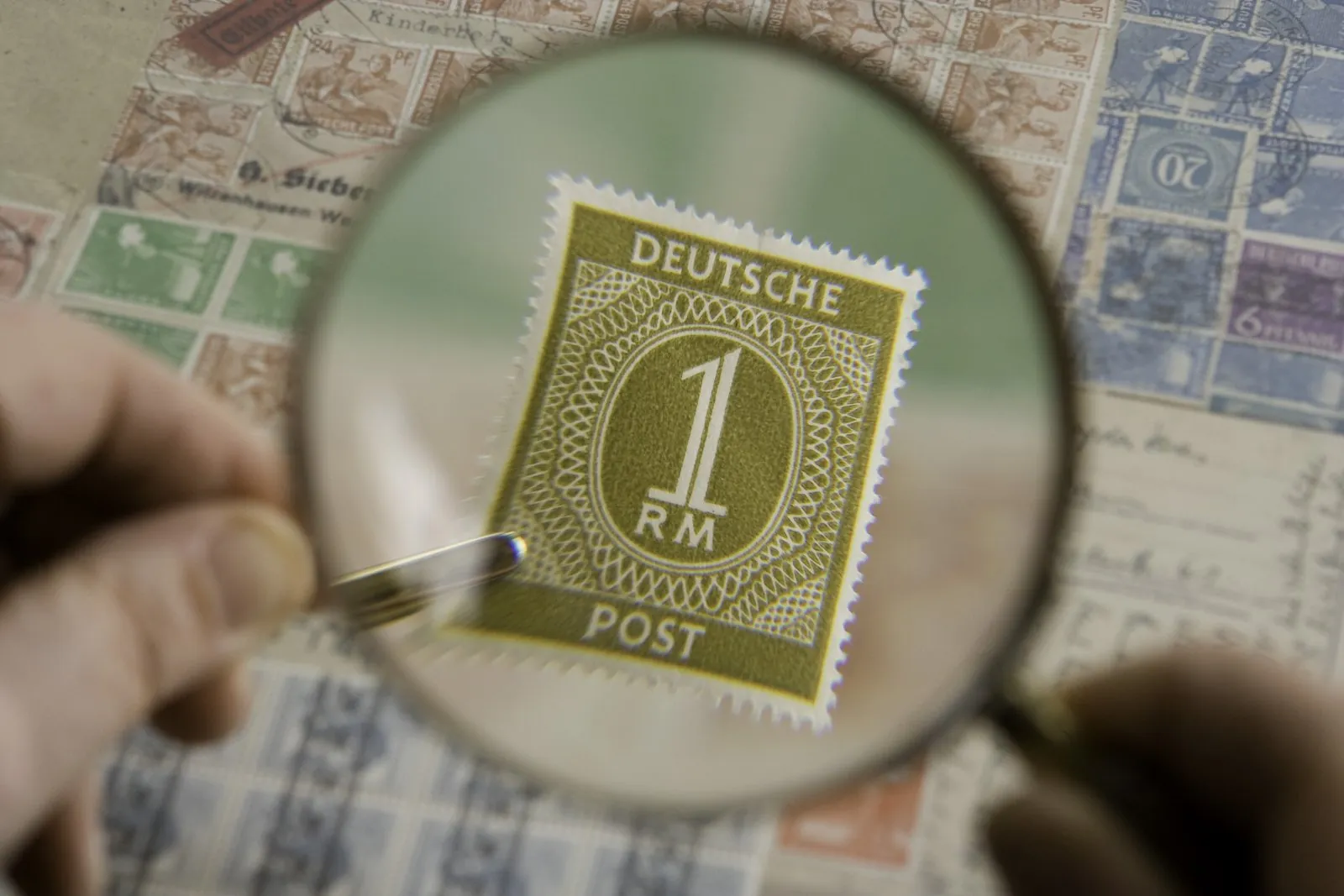 A hand holds a magnifying glass over a vintage postage stamp, enlarging its fine print