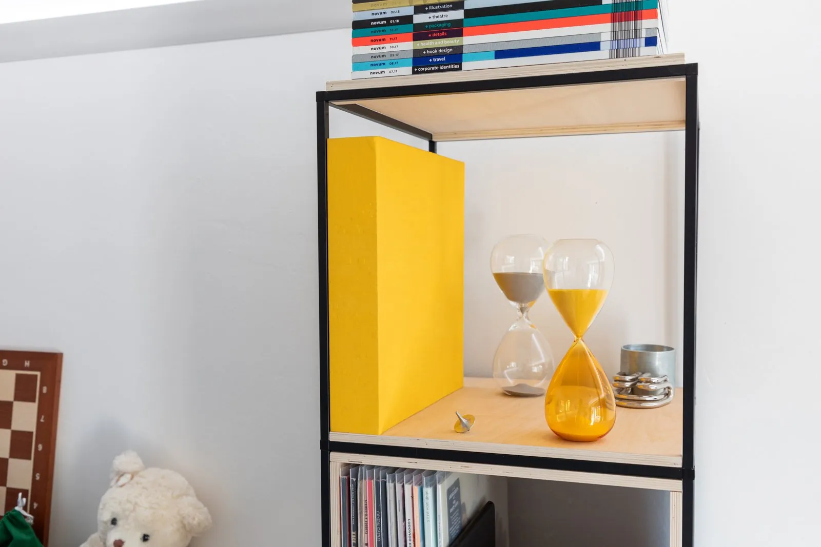 Two hourglasses, one with yellow sand and one with gray sand, on a wooden shelf next to a yellow box